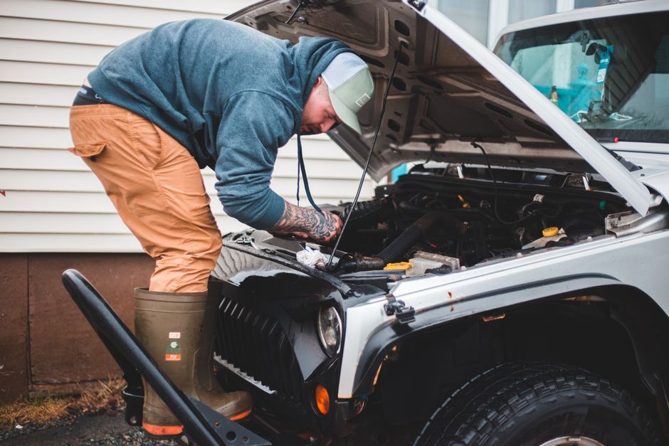 A man wearing a green cap, a blue sweatshirt, and tan trousers is leaning over the open hood of a silver off-road vehicle, inspecting or fixing the engine. He is standing on a grassy area next to a wh