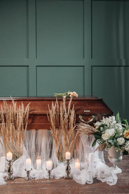 A floral arrangement featuring white roses, peonies, and greenery in a tall white vase, set on a wooden floor in front of a green paneled wall, alongside pampas grass stalks arranged in clear glass va