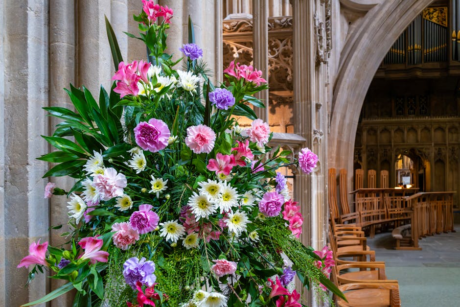 A lush floral arrangement featuring pink carnations, white daisies, purple lisianthus, and green foliage, set in a tall, dynamic bouquet held upright beside a stone pillar within St Nicholas Church in