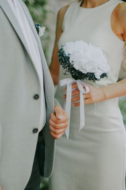 A bride holding a bouquet featuring white hydrangeas, complemented by dark green foliage and tied with a white satin ribbon, standing beside a groom dressed in a light grey suit with a white shirt, in